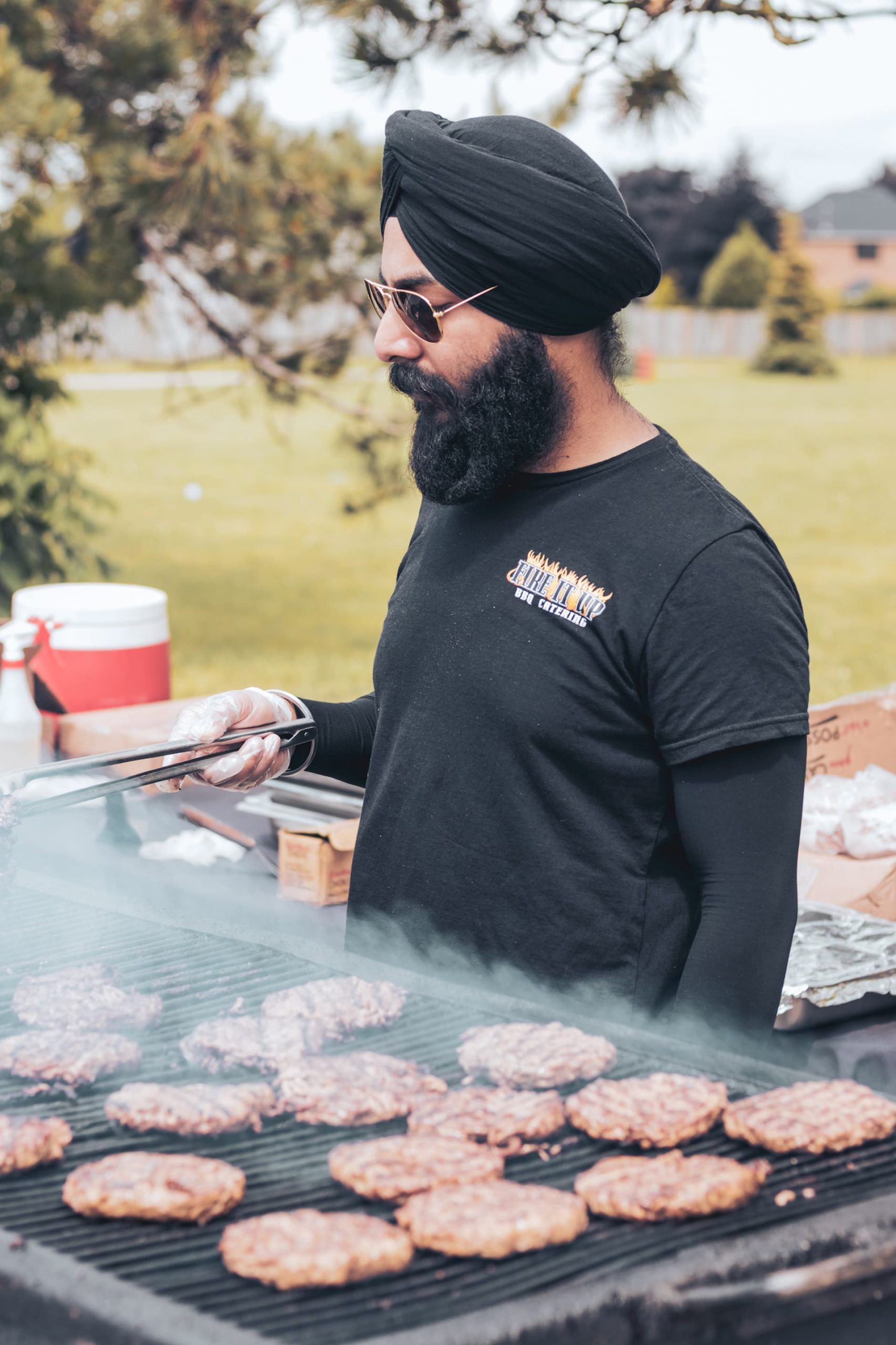 A man wearing a black turban and sunglasses grills burgers outdoors, holding tongs over a smoky barbeque.