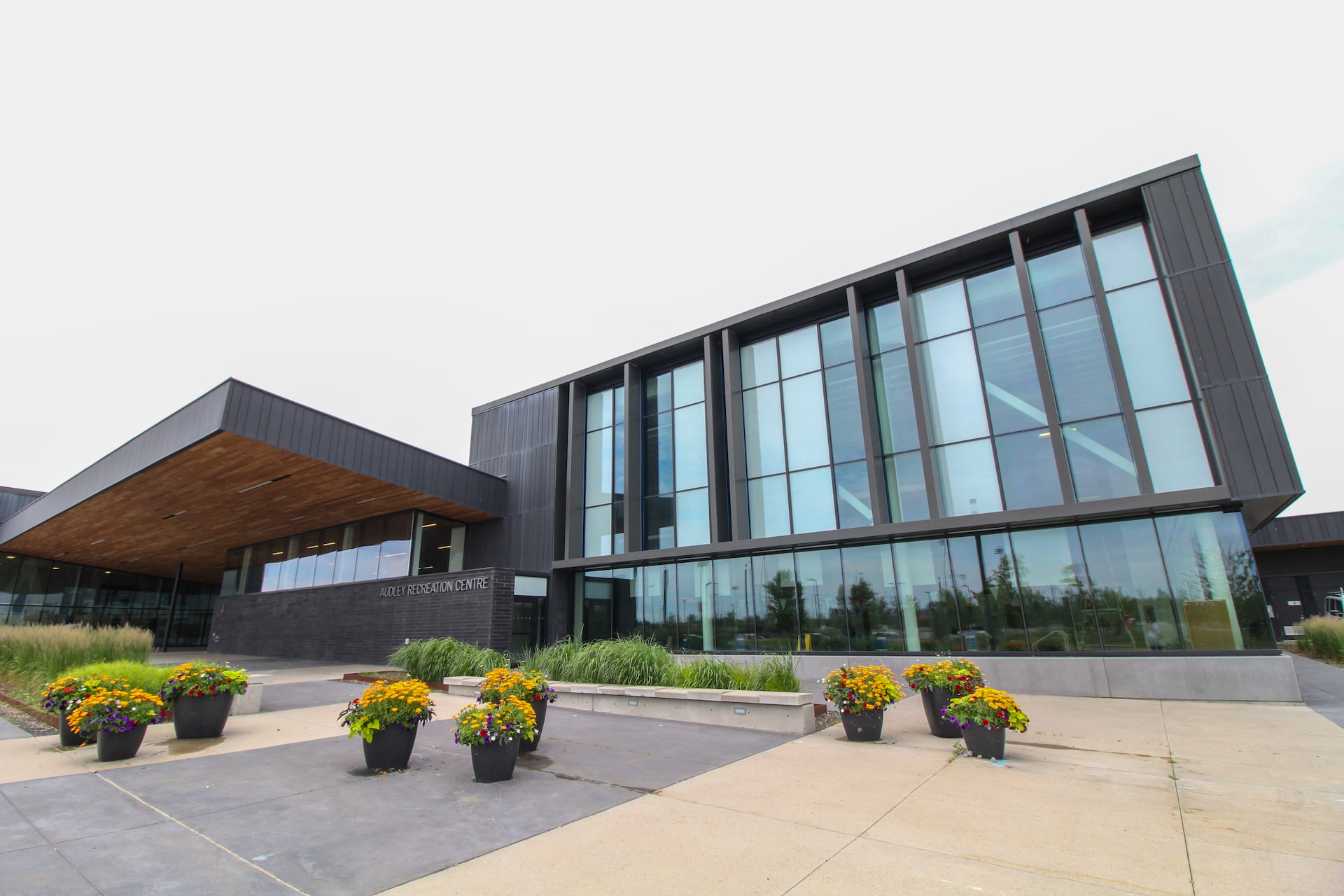 Modern glass building with large windows, landscaped planters, and a concrete walkway in front on an overcast day.