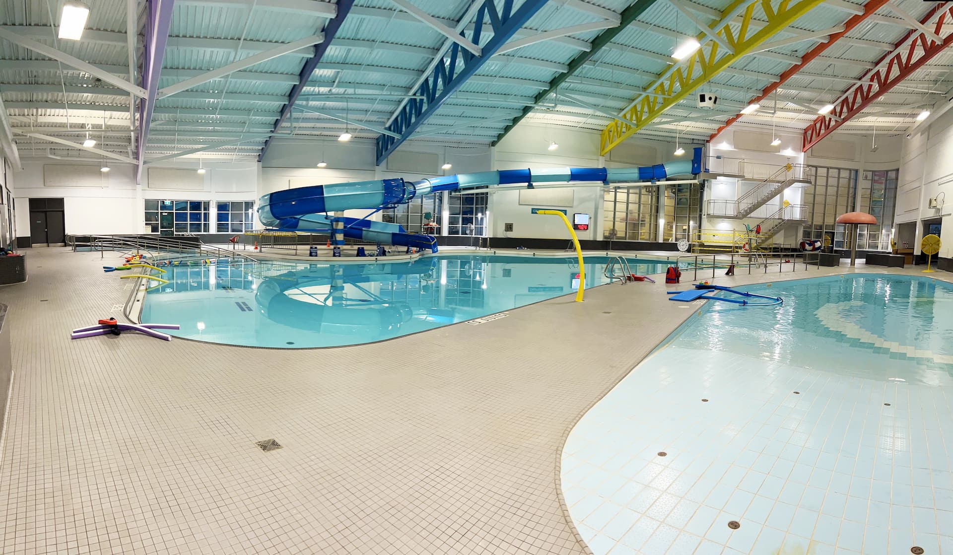 Indoor swimming pool with a blue slide, pool noodles, and colourful beams on the ceiling; no people present.