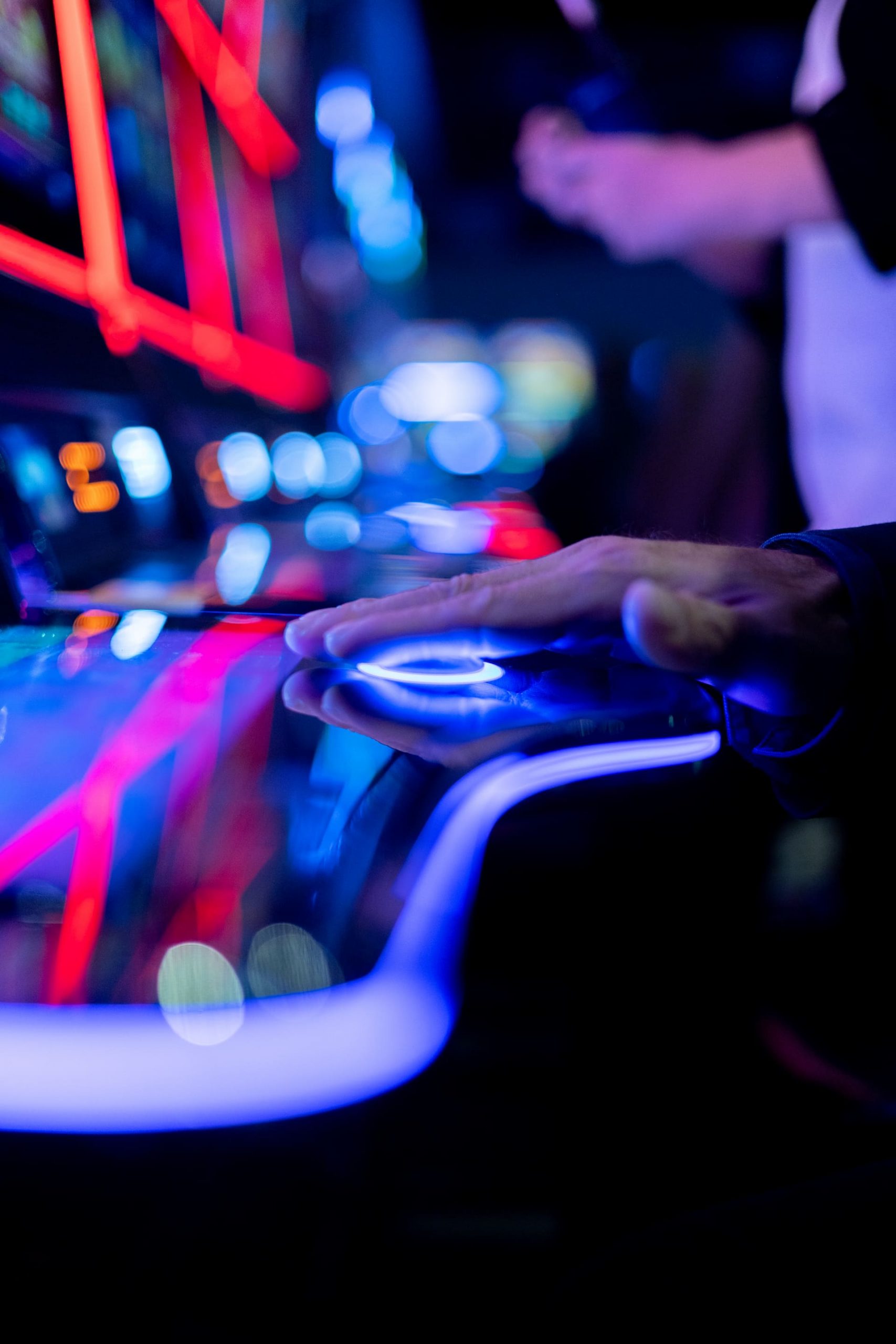 A person’s hand resting on a brightly lit fruit machine in a casino with colourful neon lights in the background.