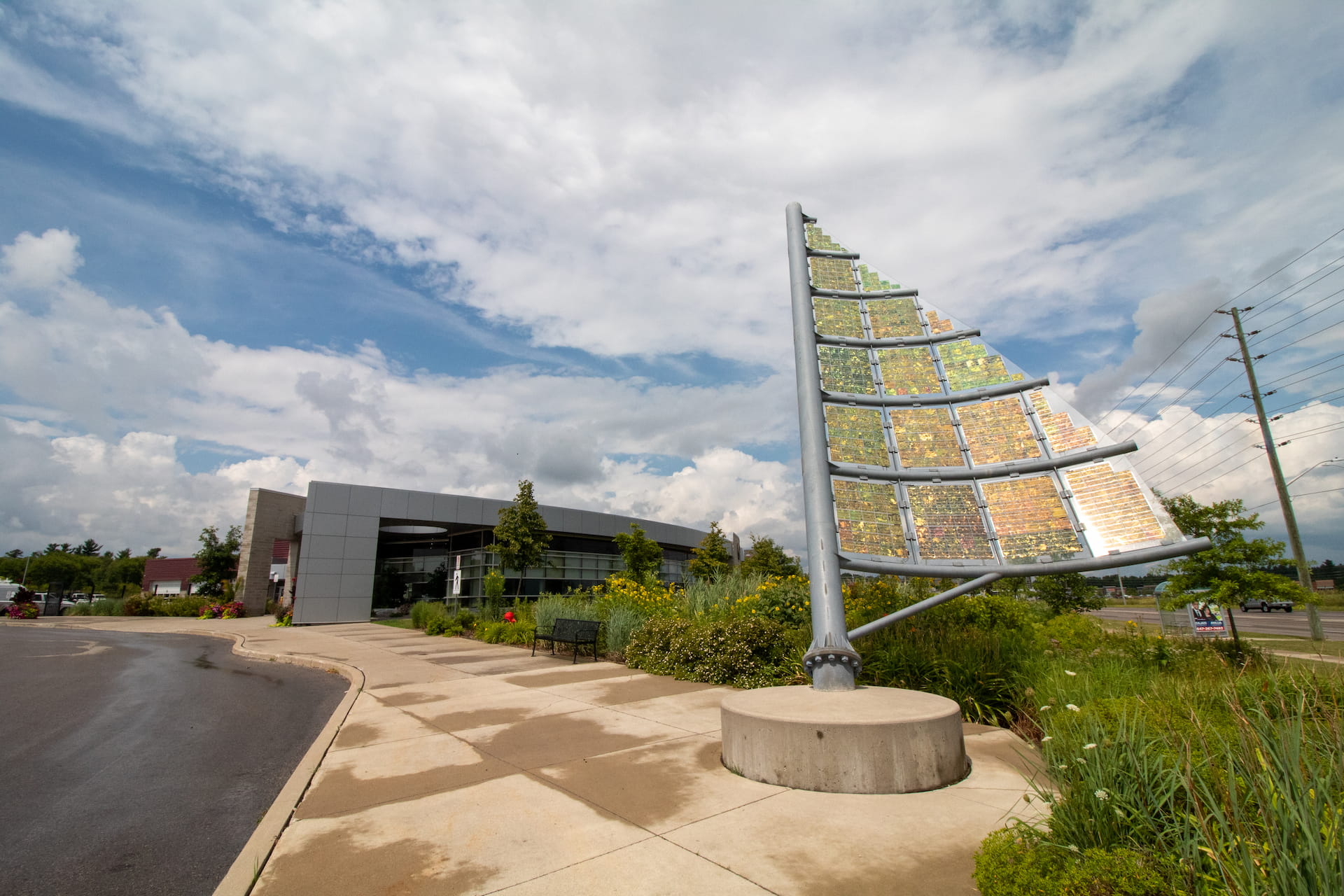 A large outdoor mosaic sculpture resembling a sail stands near a modern building and landscaped pavement.