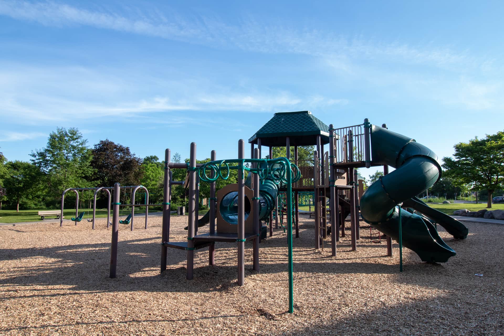 Outdoor playground with swings, climbing structures, and a spiral slide on a bark surface, surrounded by trees under blue sky.