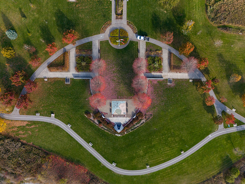 Aerial view of a landscaped park with curved paths, a central pavilion, colourful trees, and a parked white car.
