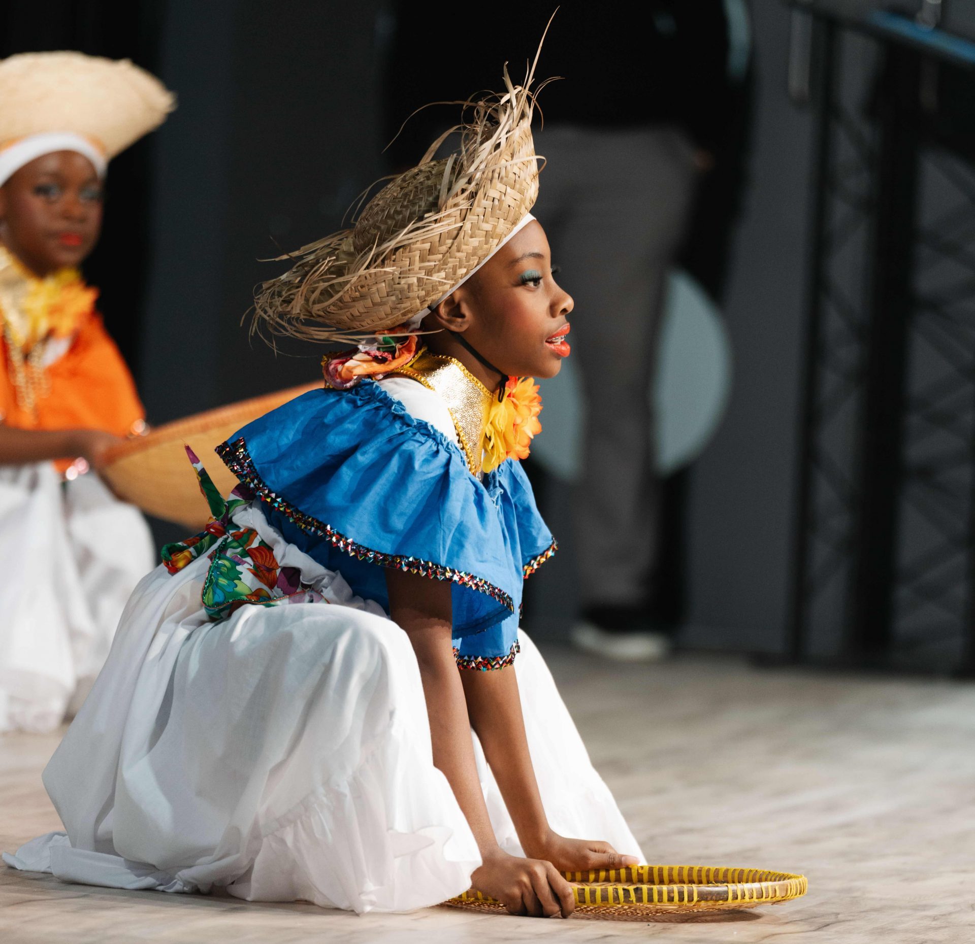 A girl in traditional attire kneels on the floor holding a woven basket, performing a cultural dance.