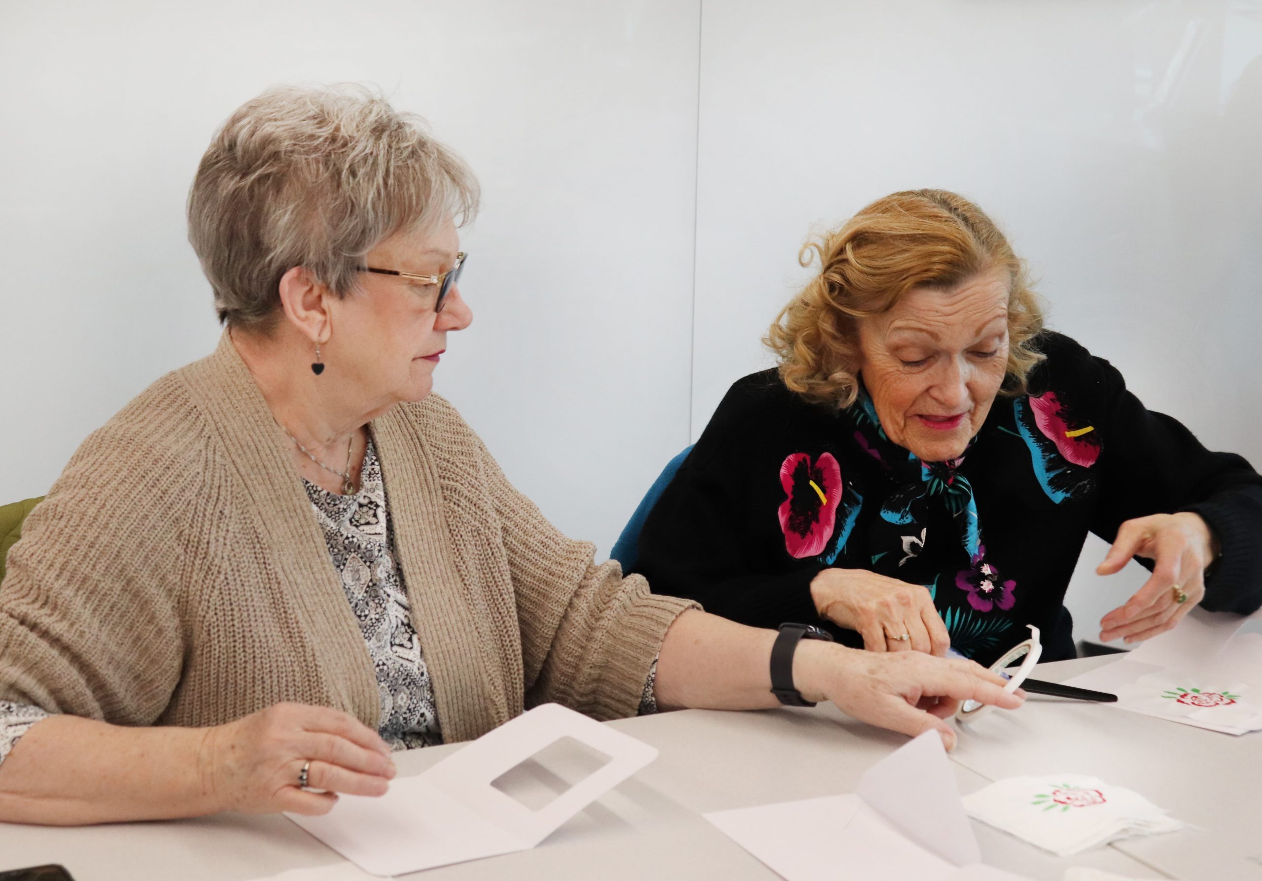 Two older women sit at a table, working together on a papercraft project with cards and cut-out shapes.