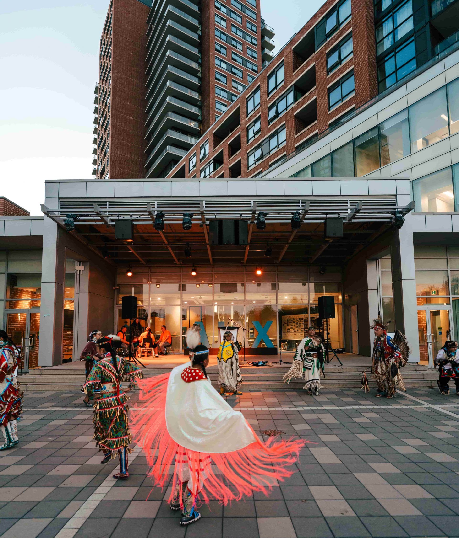 Indigenous dancers perform in traditional regalia outside a modern building on a paved piazza.