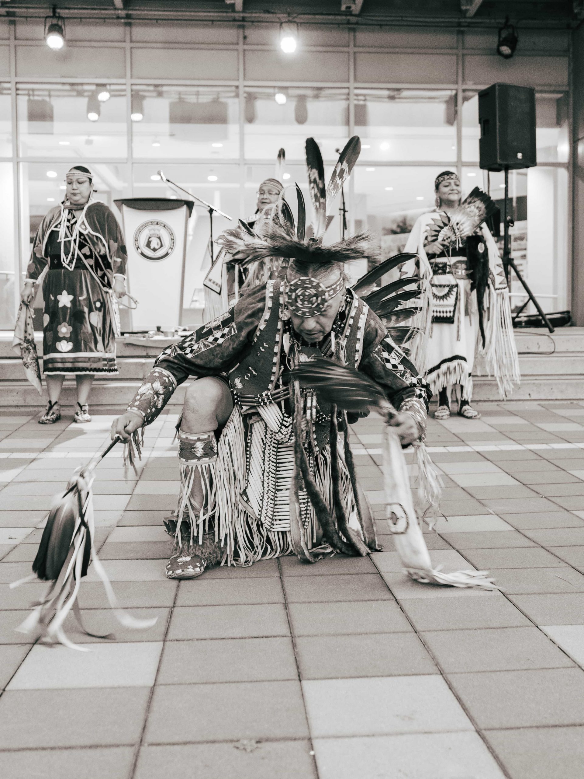 A group of Indigenous dancers in traditional regalia perform on an outdoor tiled plaza in front of a building.