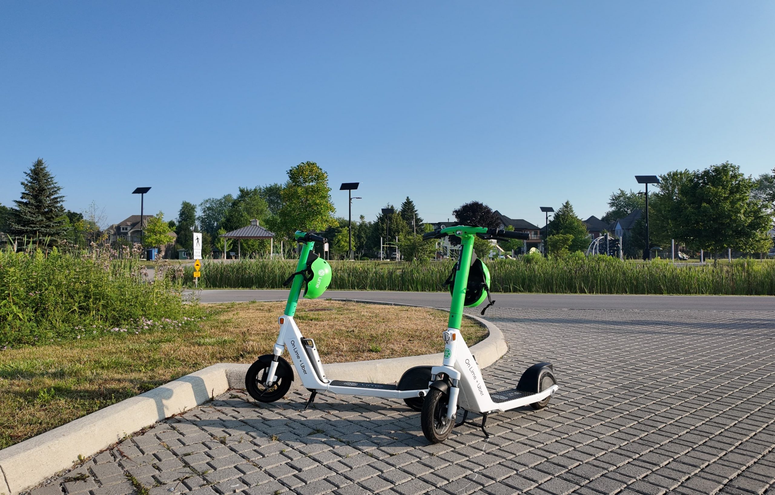 Two electric scooters parked on a paved path near a grassy area and residential houses under a clear blue sky.