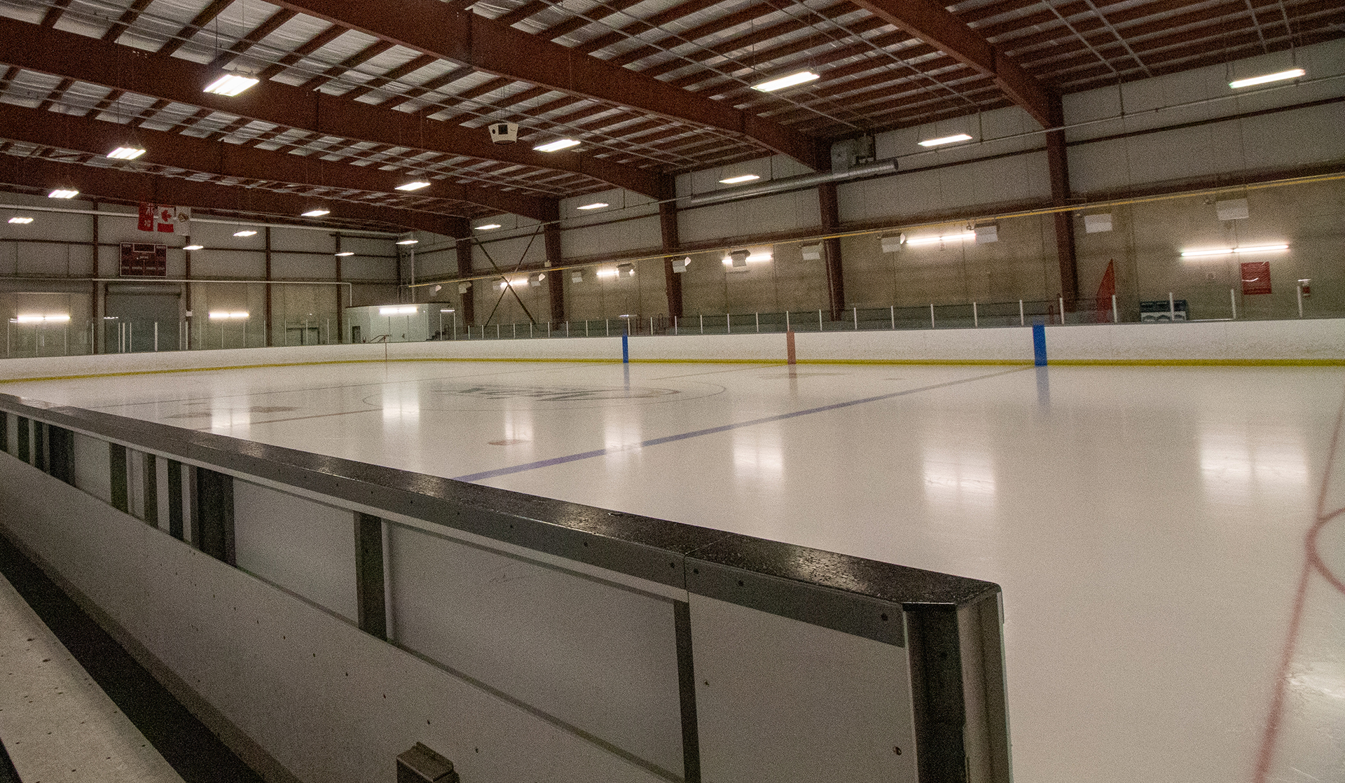 Empty indoor ice hockey rink with clear boards, overhead lights, and visible blue and red lines on the ice.