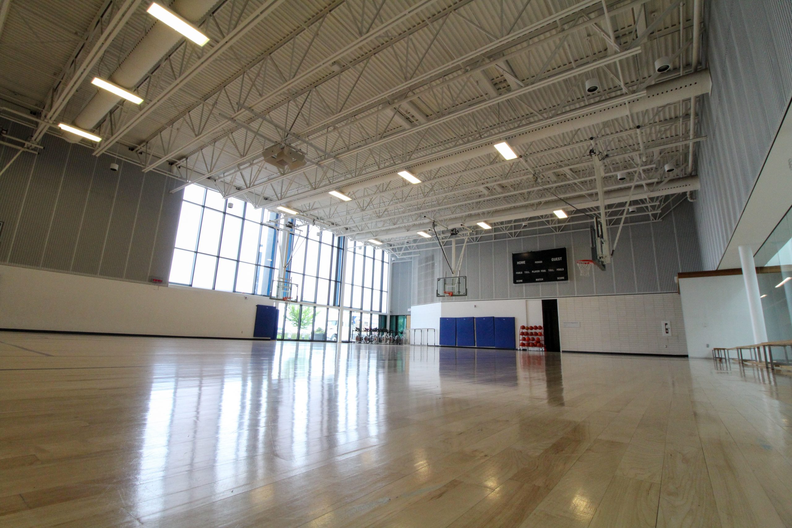 Empty indoor basketball court with polished wooden floor, high ceiling, basketball hoops, and large windows.