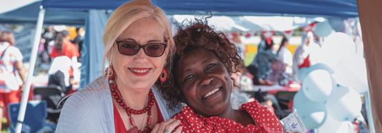 Two women smiling and posing together at an outdoor event with marquees and balloons in the background.