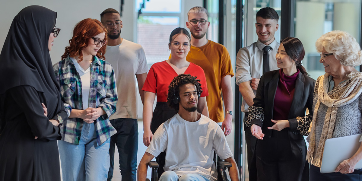 A diverse group of people, including one person in a wheelchair, gathers and interacts in a bright indoor setting.