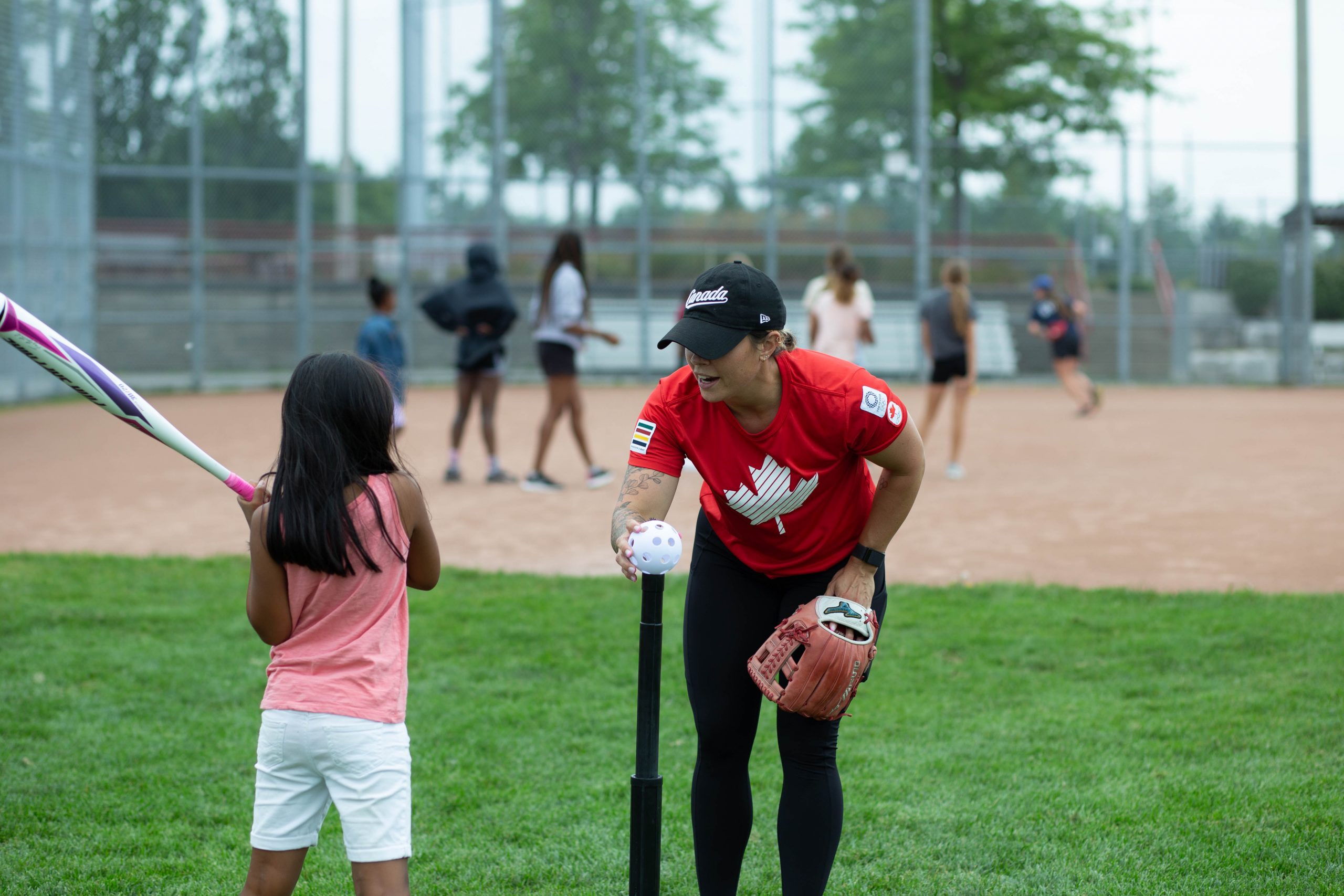 A coach helps a young girl prepare to hit a ball on a tee during a youth baseball practice on a pitch.