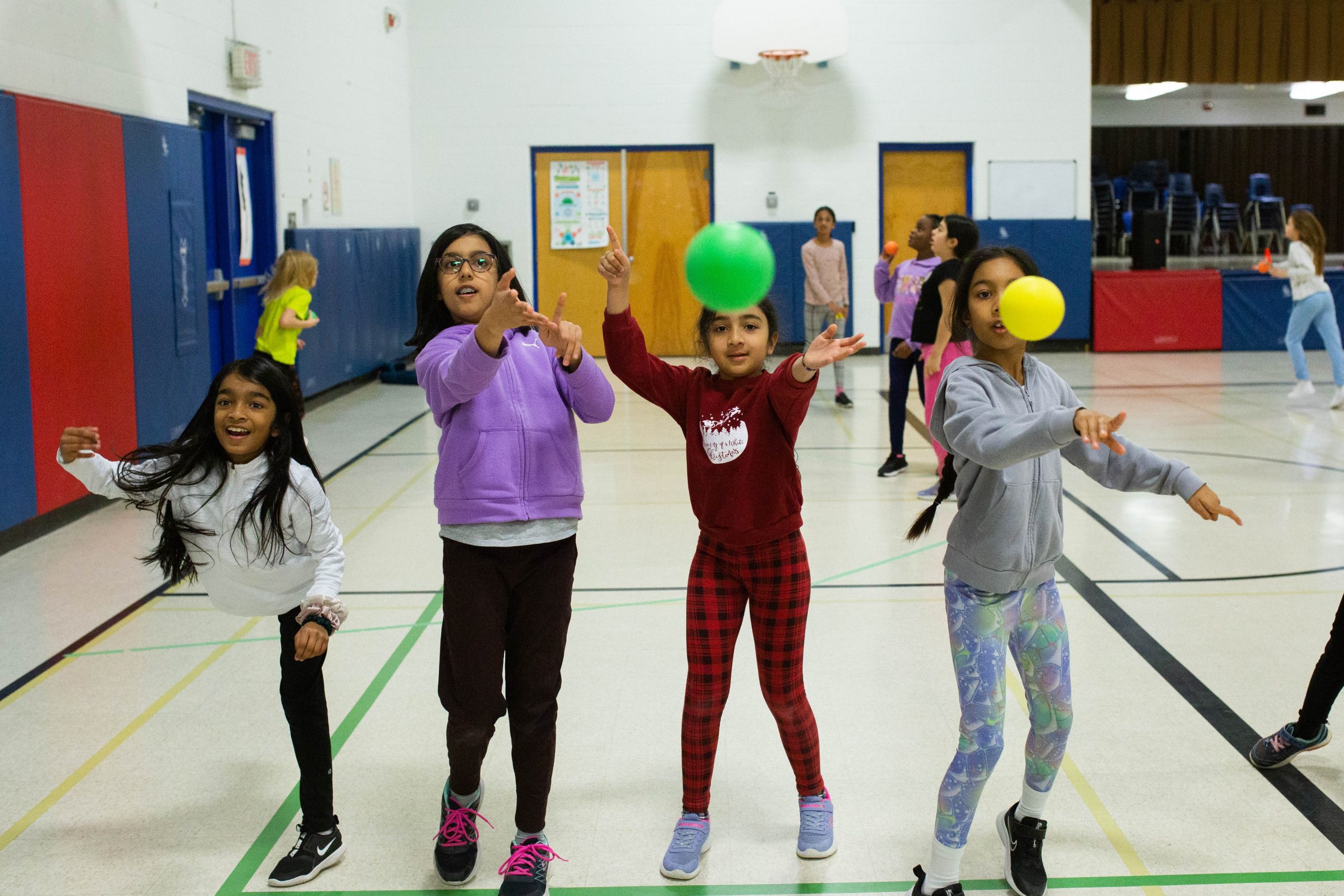 Four girls throw balls in a gymnasium whilst other children play in the background.