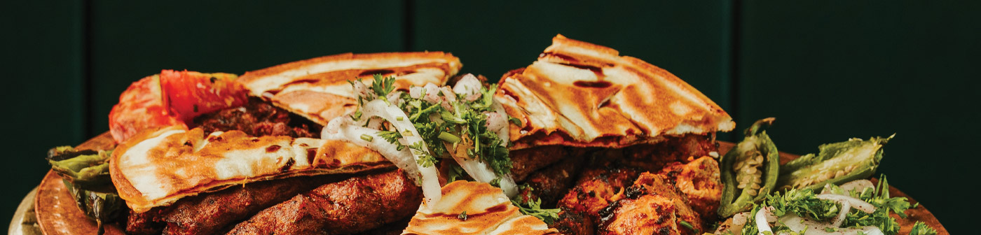 A platter with grilled meats, pitta bread, onions, and green peppers, arranged on a wooden board against a dark background.