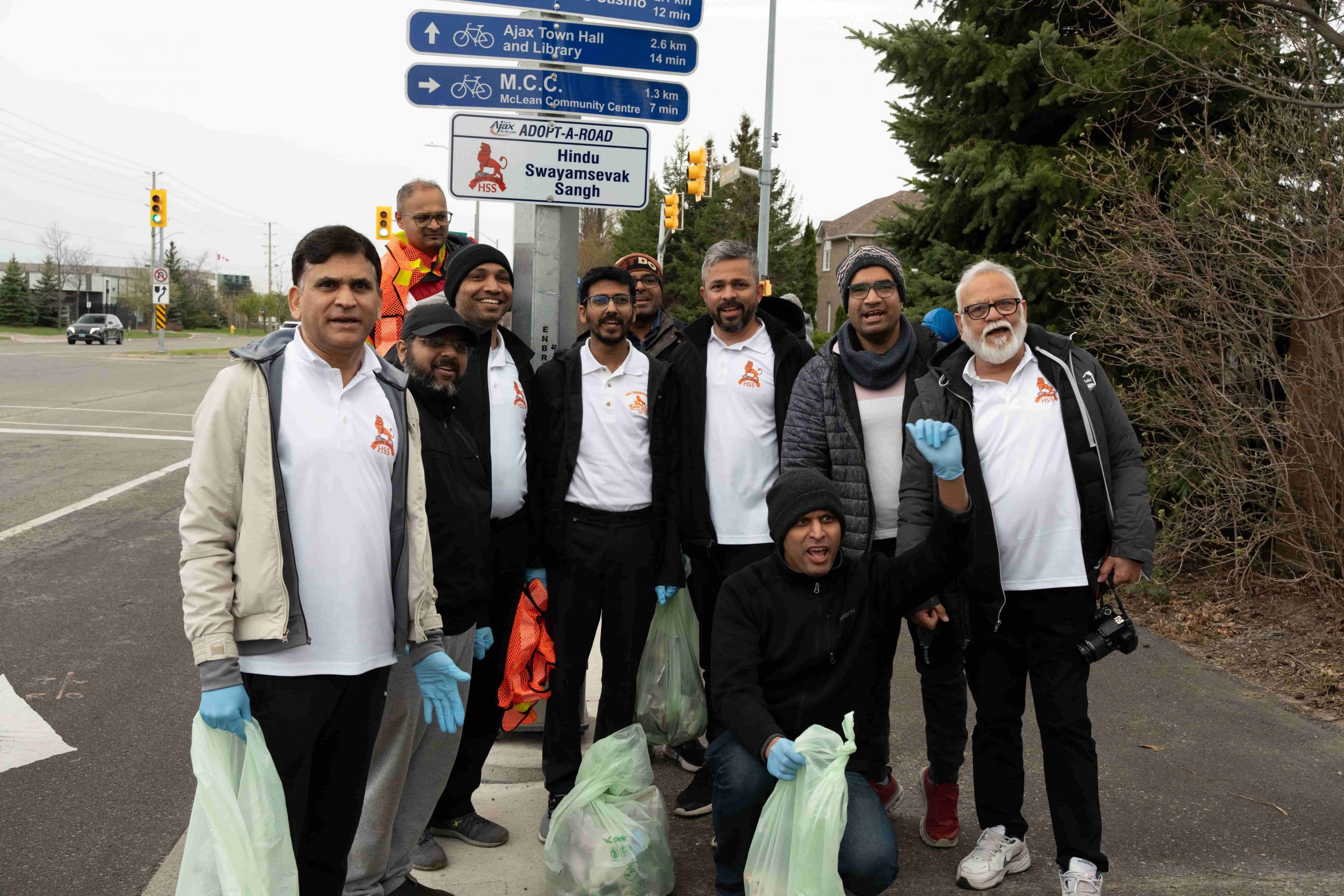 A group of men pose on a pavement holding bin bags and wearing gloves near a road sign.