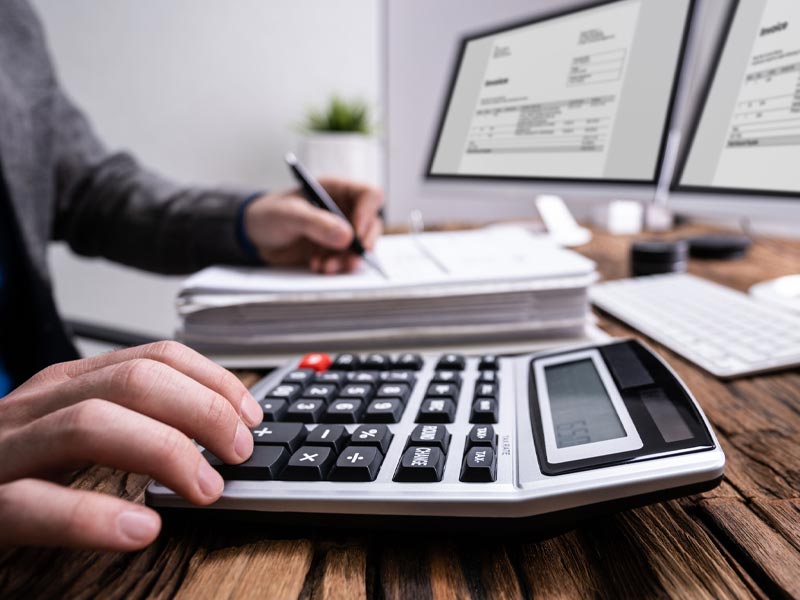 Person using a calculator and writing on documents at a desk with two computer monitors displaying spreadsheets.