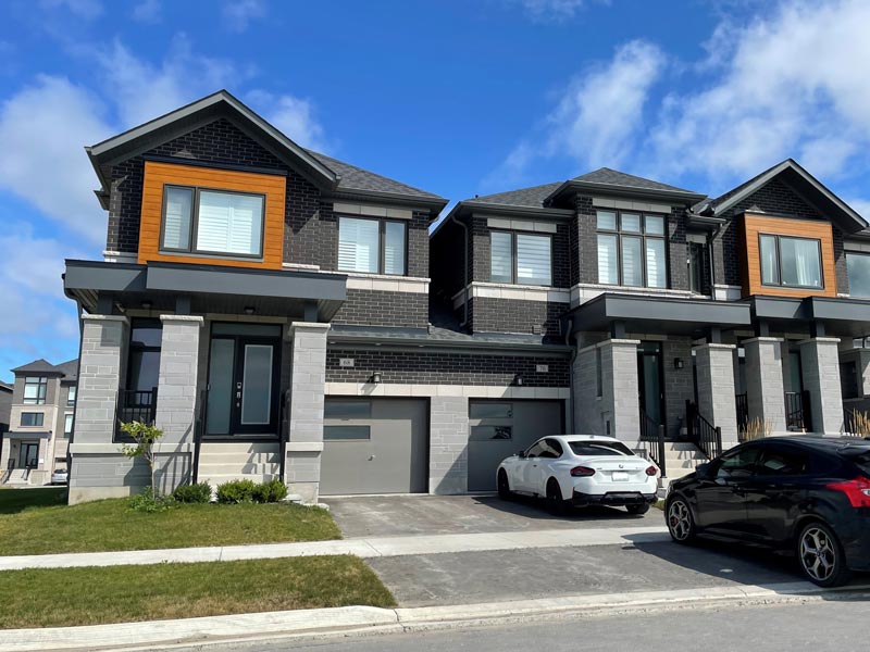 Modern two-storey duplex homes with brick and timber accents, driveway, and parked cars under a blue sky with clouds.