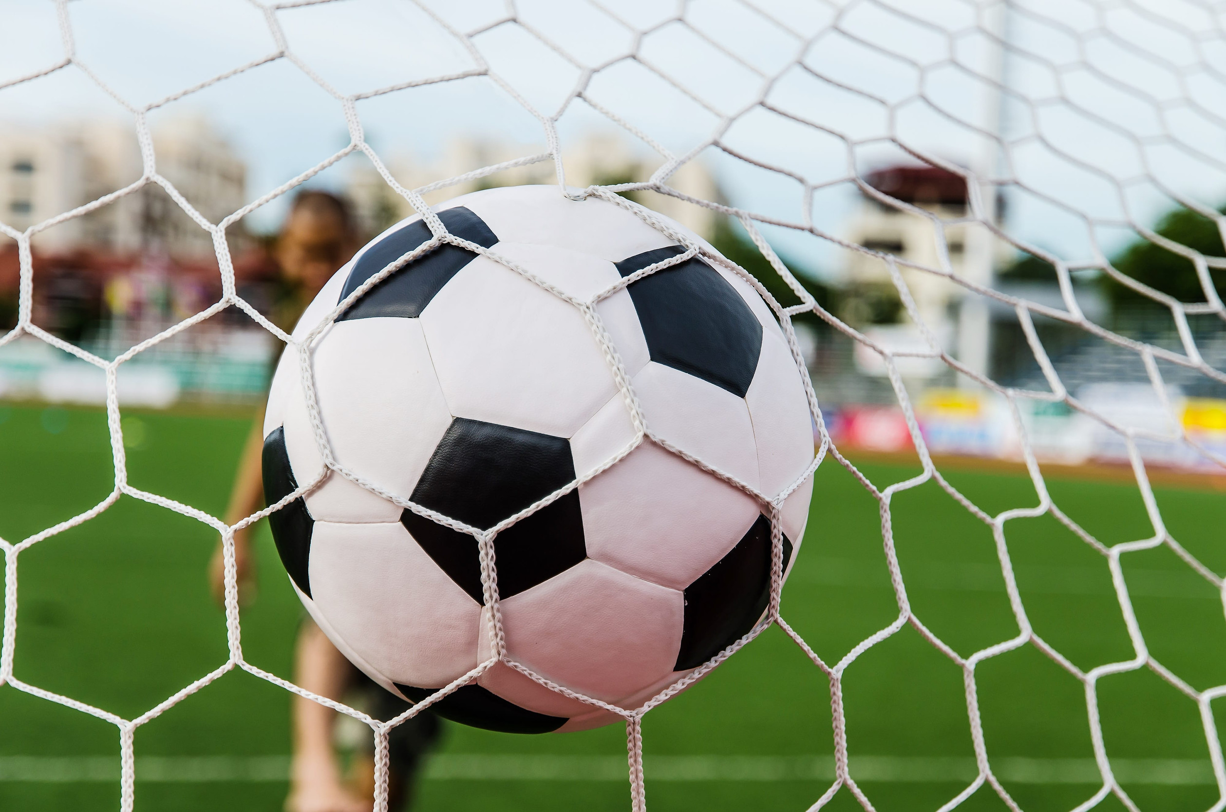 A football caught in the net of a goal on a green pitch, with blurred stadium seats in the background.