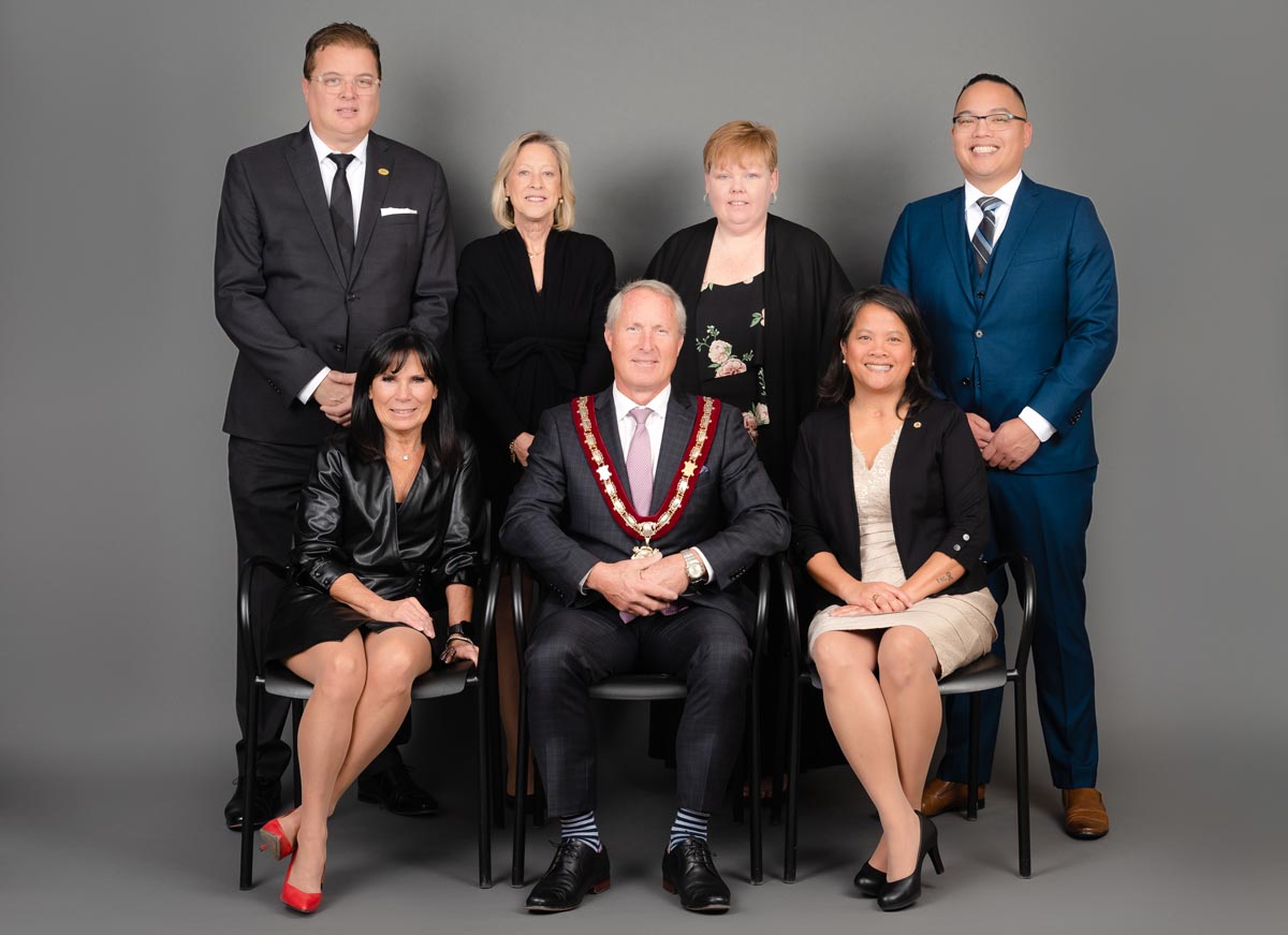 Seven formally dressed adults pose for a group portrait in front of a grey background; one wears a ceremonial chain.
