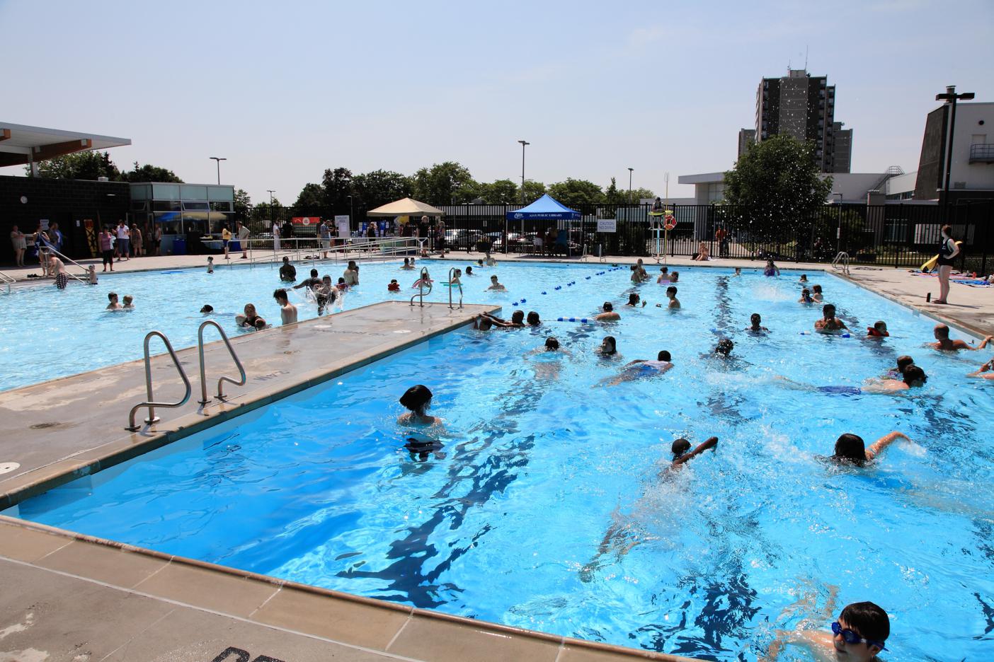 Outdoor public swimming pool with many people swimming and playing in separate deep and shallow sections on a sunny day.