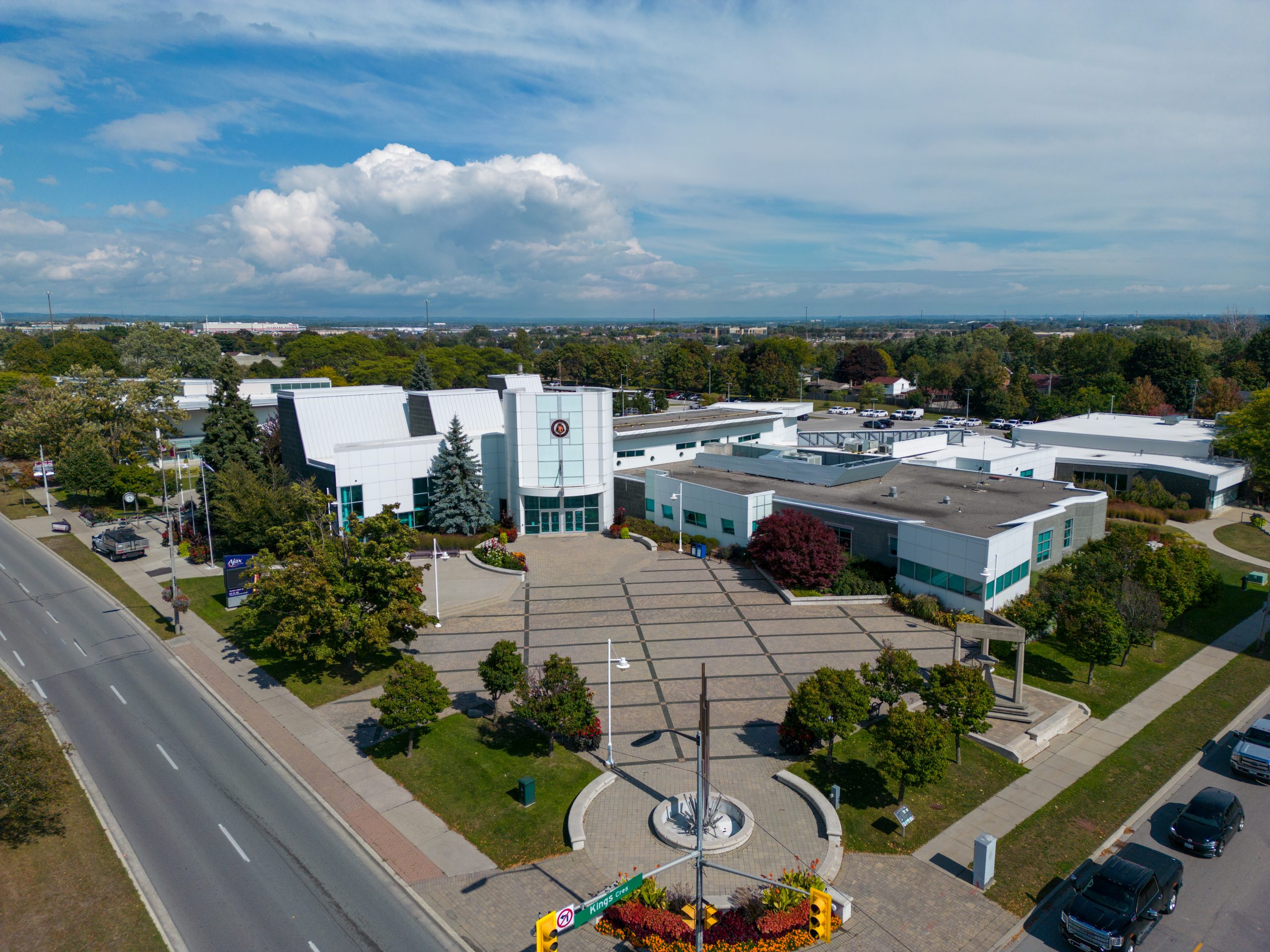 Aerial view of a modern white building complex with a landscaped entrance and adjacent road on a clear day.