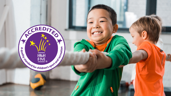 Two children smiling whilst playing tug-of-war indoors, with a "High Five Accredited" logo for children's sport and recreation.