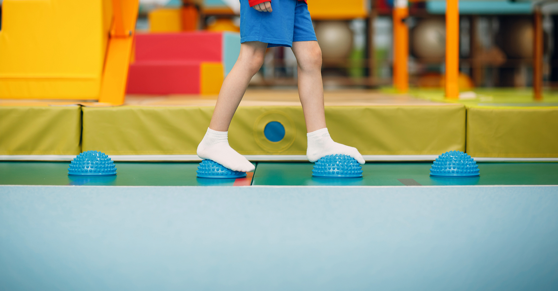 Child in white socks balancing on blue textured half-balls in a colourful indoor play area or gym.