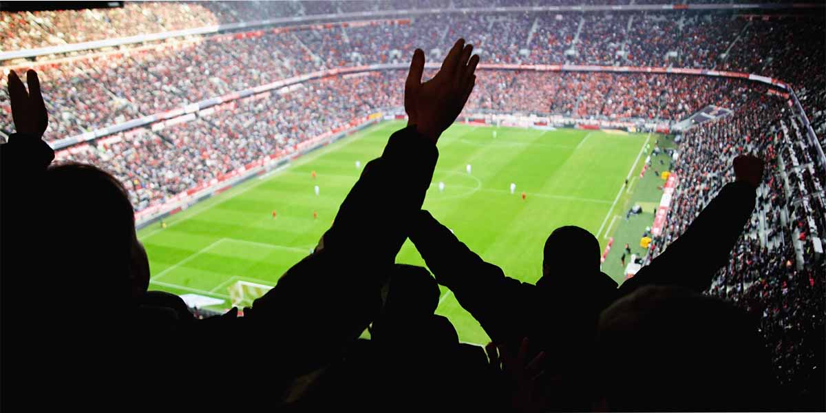 Crowd of fans with raised arms watching a football match in a large, crowded stadium.