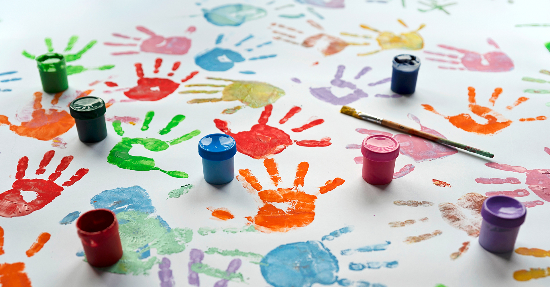 Colourful handprints cover a white surface, surrounded by open paint containers and a paintbrush.