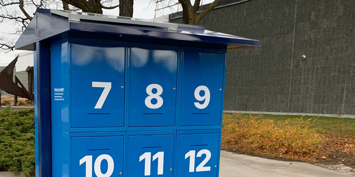 Blue outdoor locker unit with numbered doors 7 to 12, located on a pavement near a building and some bushes.