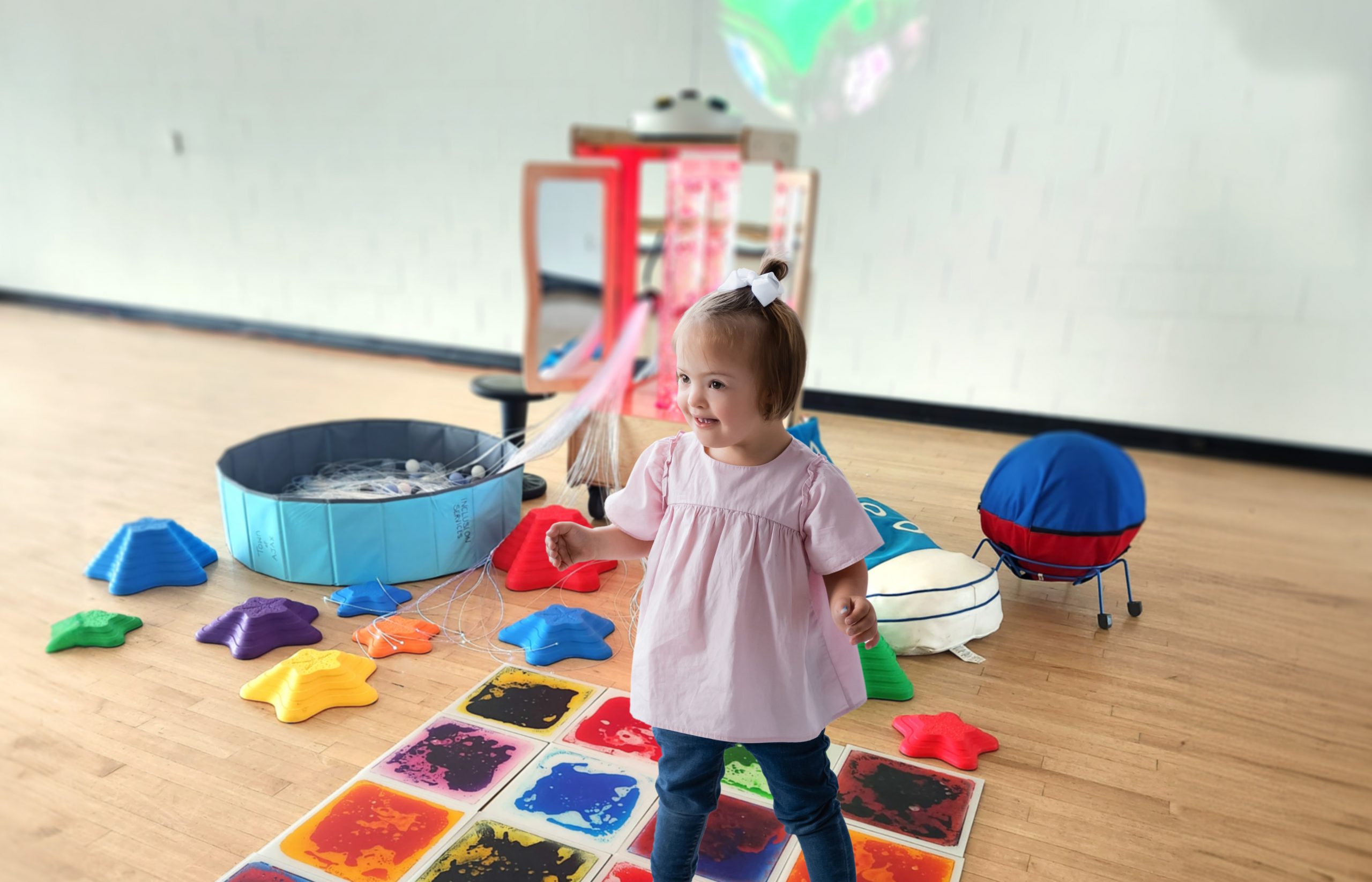 A young child stands on a colourful sensory mat in a playroom with various tactile and interactive toys.