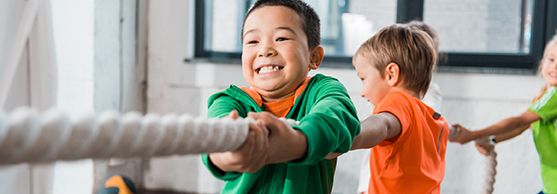 Children playing tug-of-war indoors, smiling and pulling on a thick rope together.