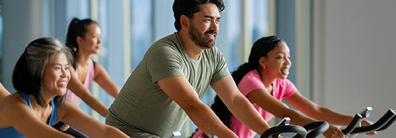 A group of adults exercise on stationary bikes in a gym, facing forwards and appearing focused.