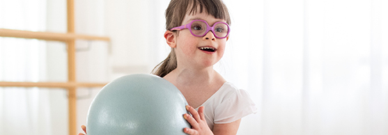 Girl with glasses holding an exercise ball in a bright room, smiling and looking forward.