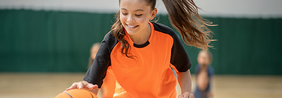 Girl in orange kit dribbling a basketball on an indoor court with a green divider in the background.