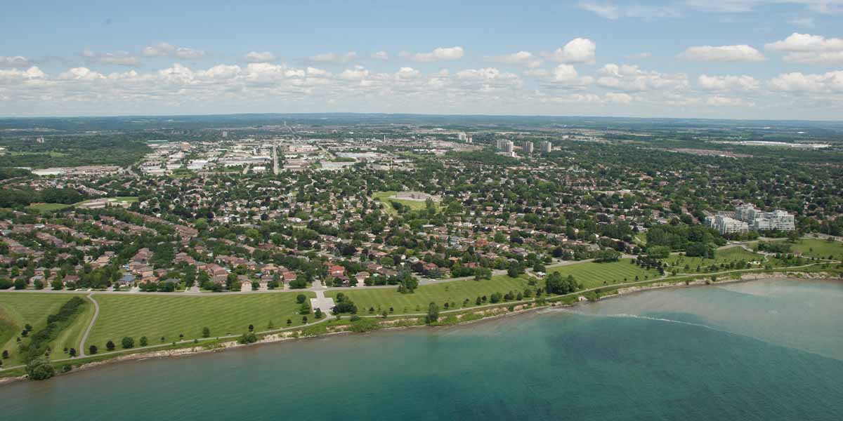 Aerial view of a suburban neighbourhood bordered by a green park and waterfront, with city buildings visible in the distance.