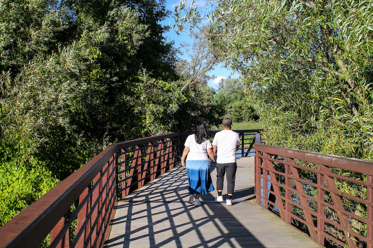 Two people walk hand in hand on a wooden bridge surrounded by green trees on a sunny day.