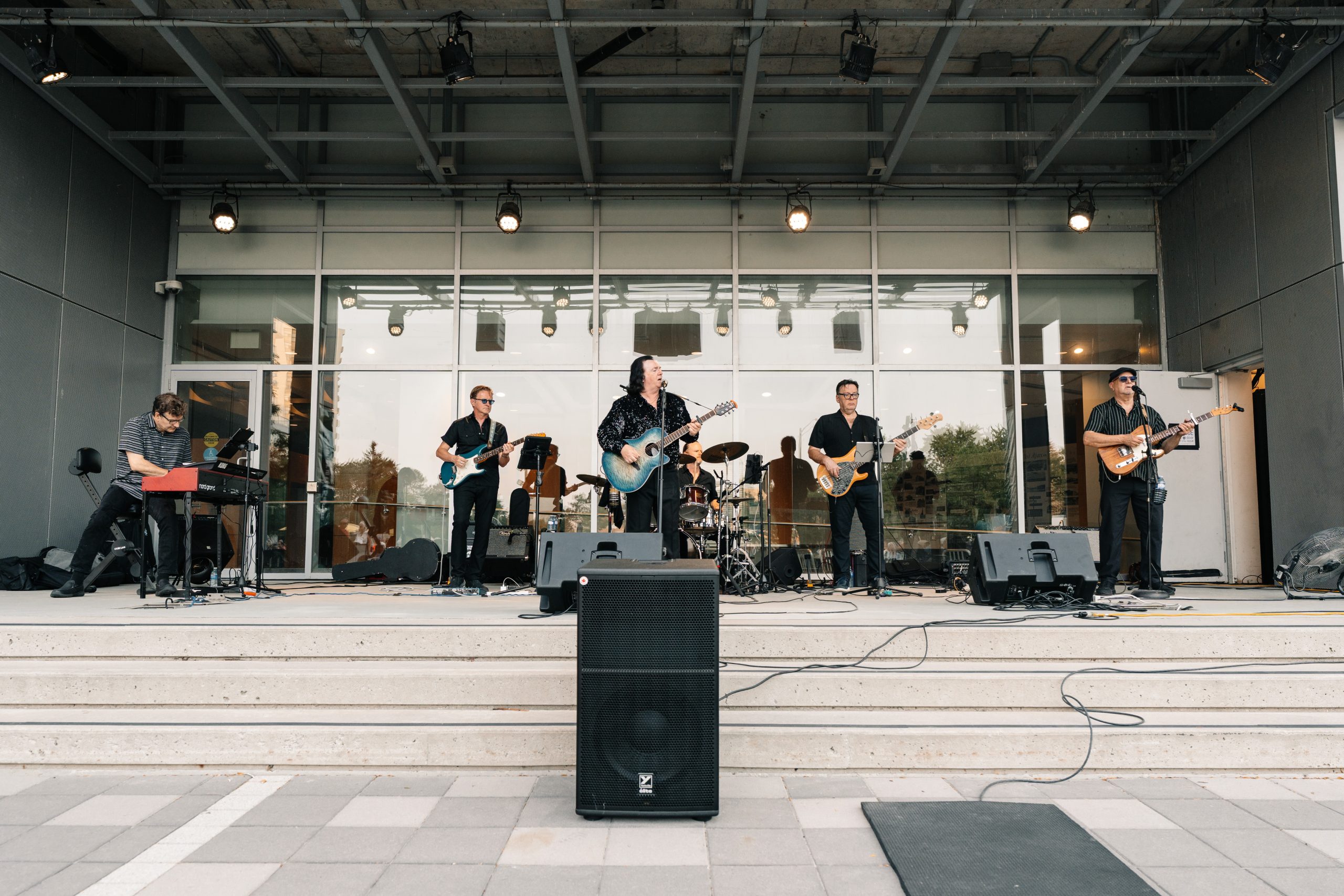 A band of six musicians performs on an outdoor stage with instruments including guitars, keyboard, and drums.