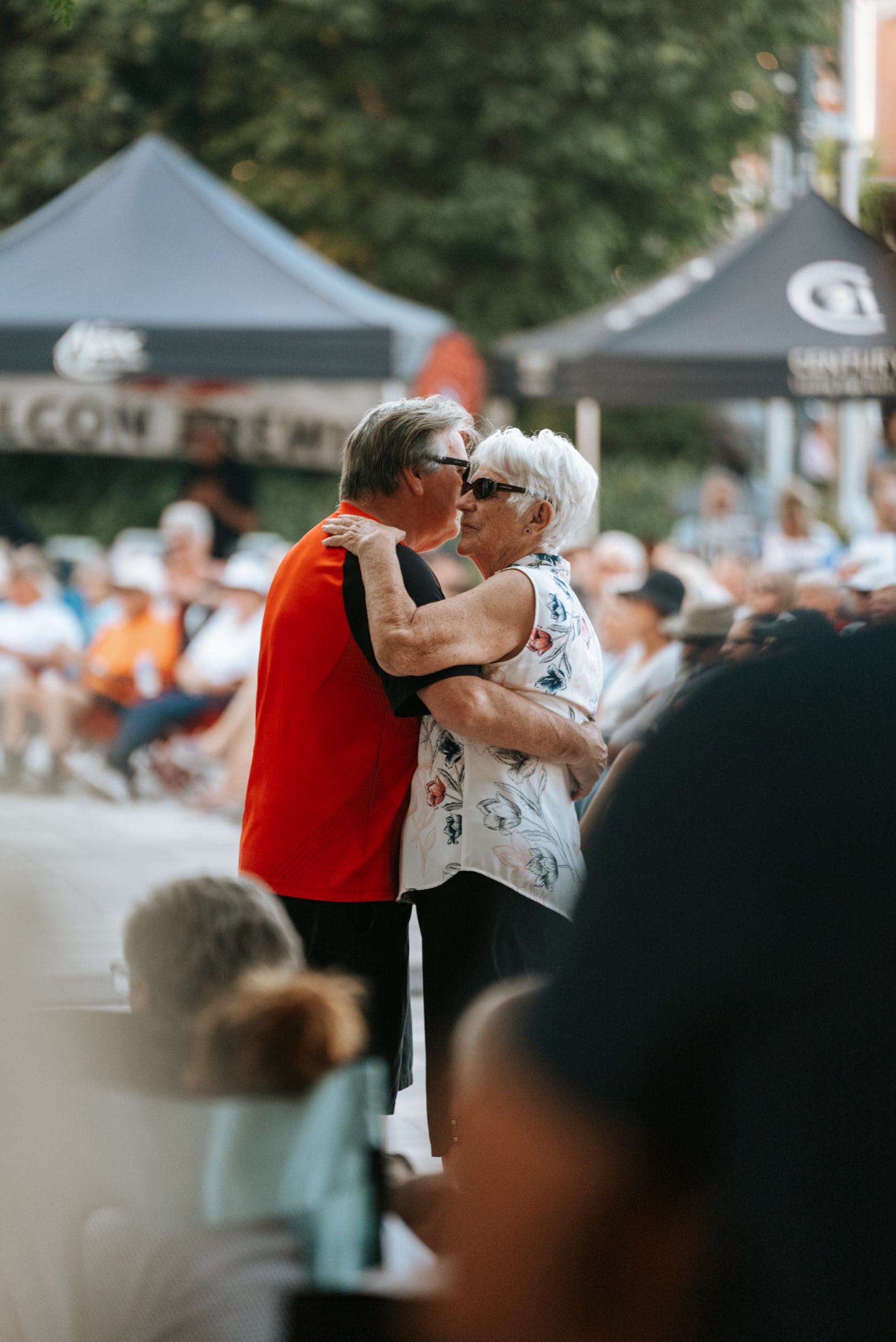 Two older adults dance together outdoors in front of a seated crowd and black event marquees.