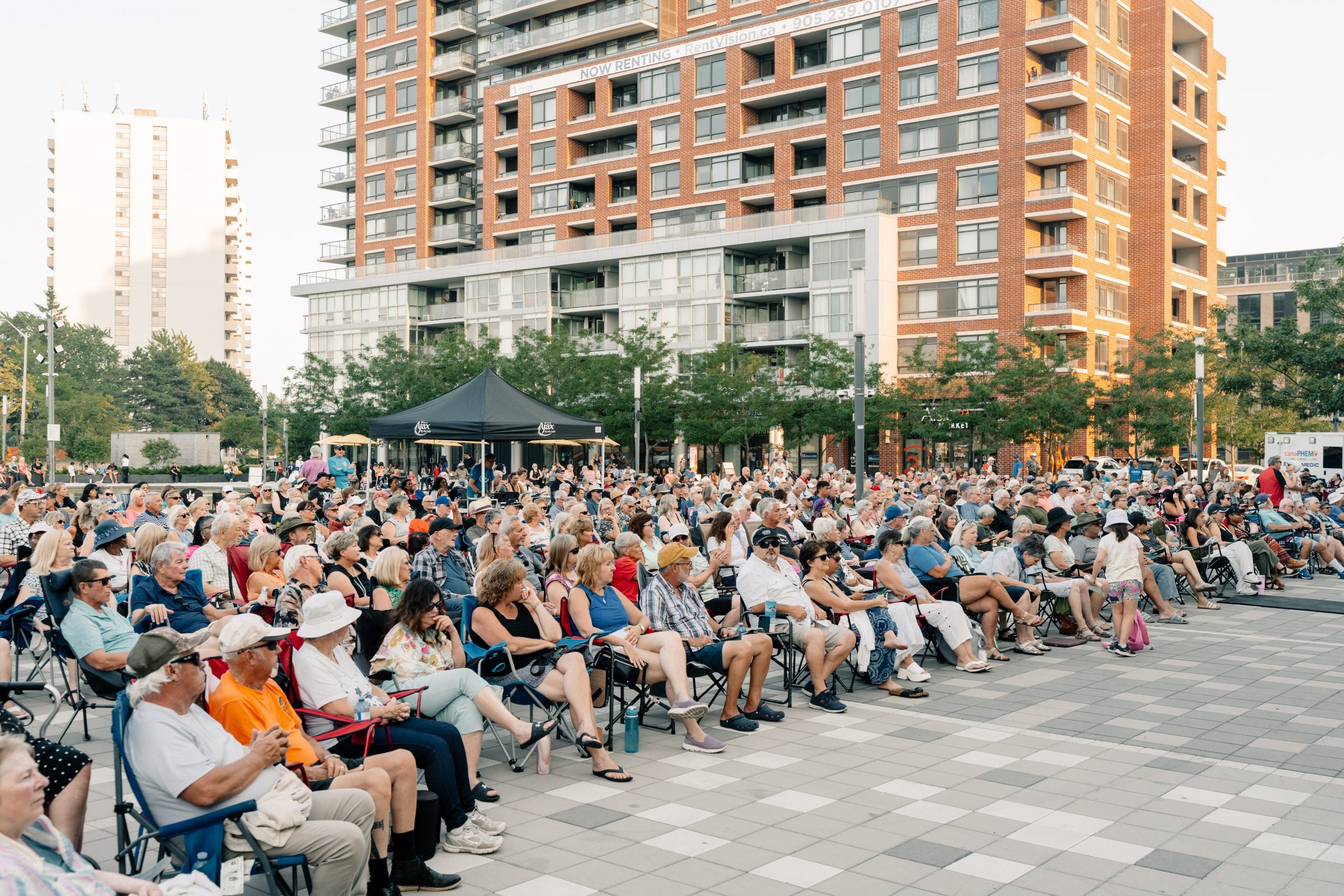 A large crowd sits outdoors in folding chairs facing a stage, with tall buildings in the background.