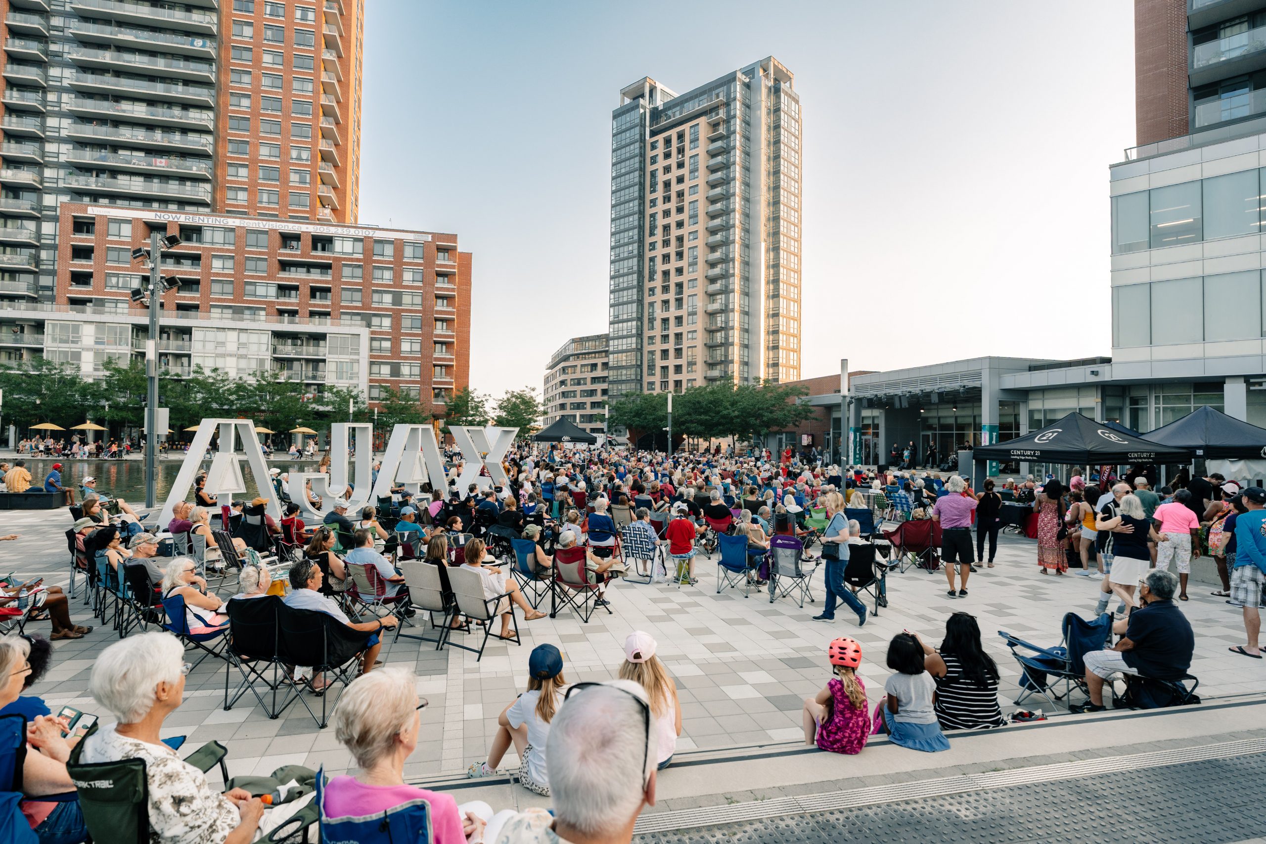 A large crowd sits and stands in a city plaza with "AJAX" letters and tall buildings in the background.