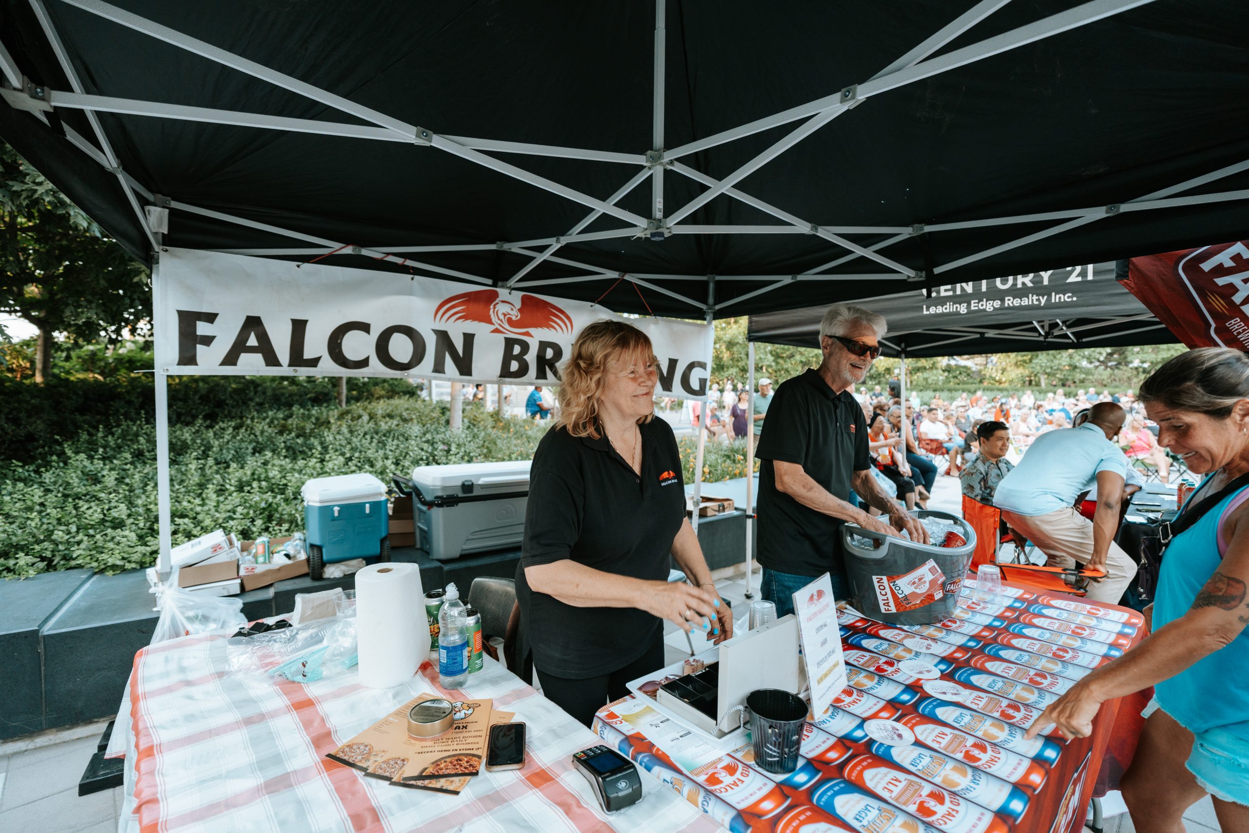 Two people stand behind a stall labelled "Falcon Brewing," handing out items at an outdoor event.