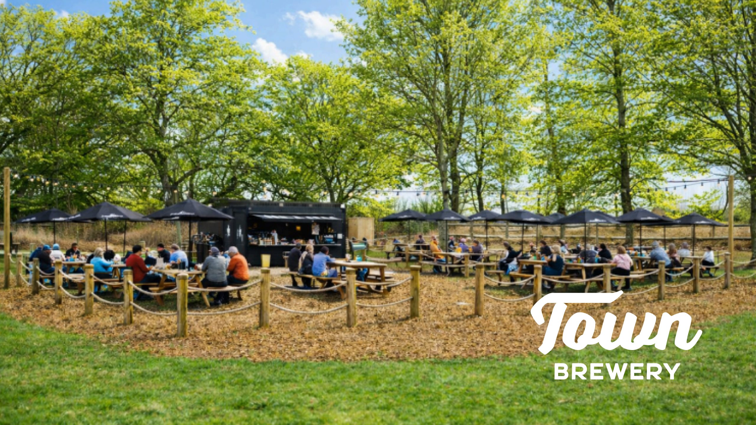 Outdoor brewery patio with people seated at picnic tables under trees, with a central bar and black umbrellas. "Town Brewery" text.