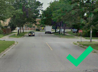 A car turns left at a junction while another vehicle waits at a give way sign; trees line both sides of the road.
