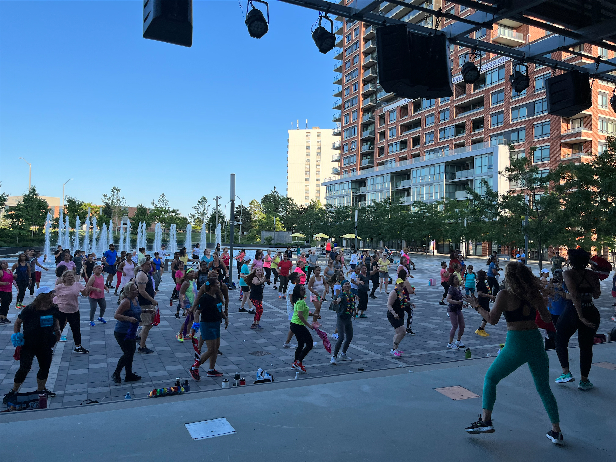 A group fitness class takes place outdoors in a city square near fountains, with participants following an instructor.