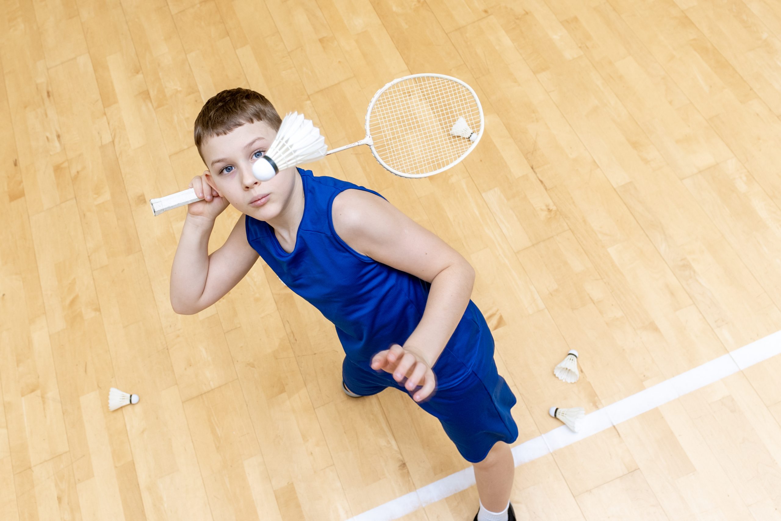 Boy in blue sportswear playing badminton indoors, holding a racquet with a shuttlecock balanced near his eye.