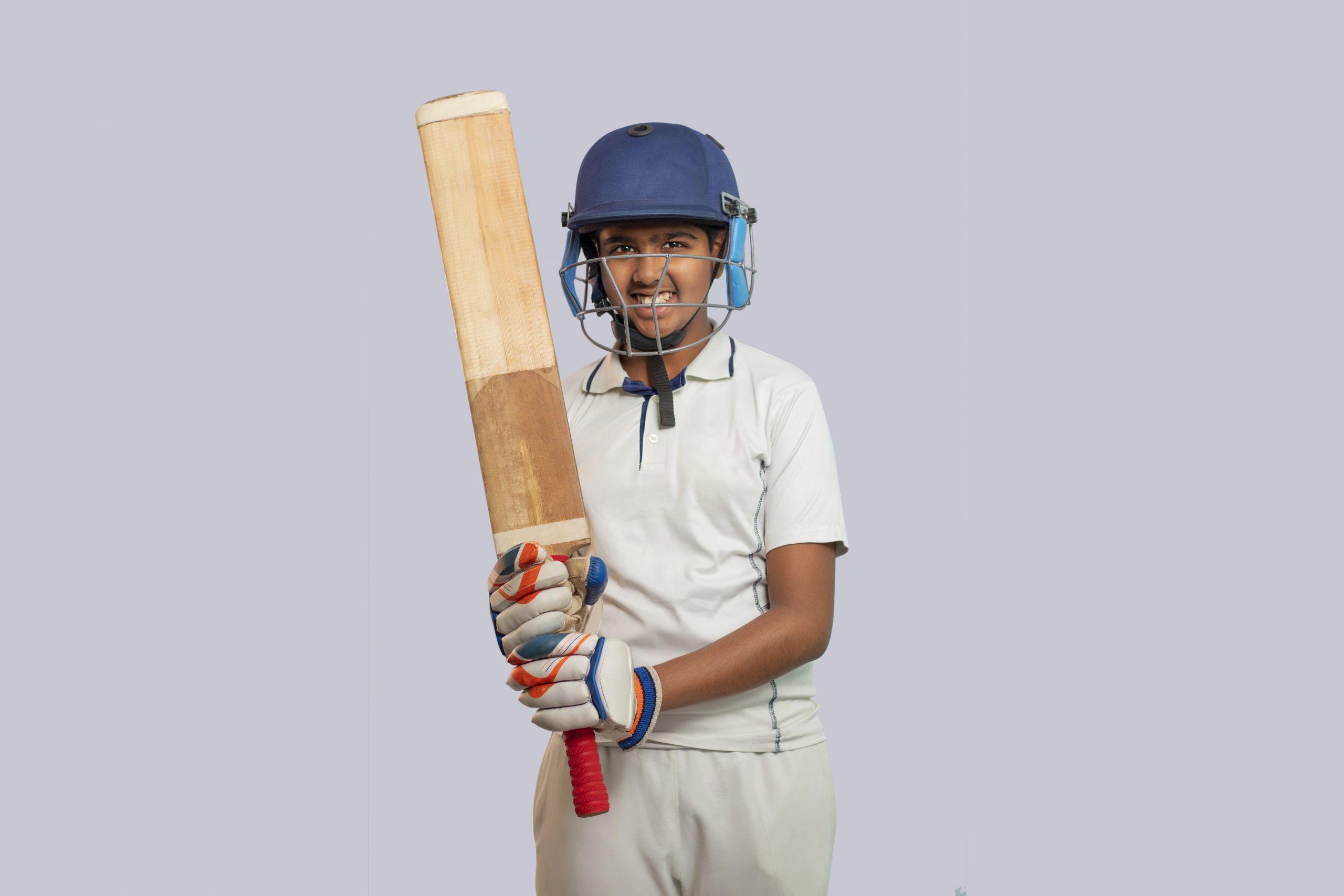 Young cricket player in full kit holding a bat, standing against a plain light background.