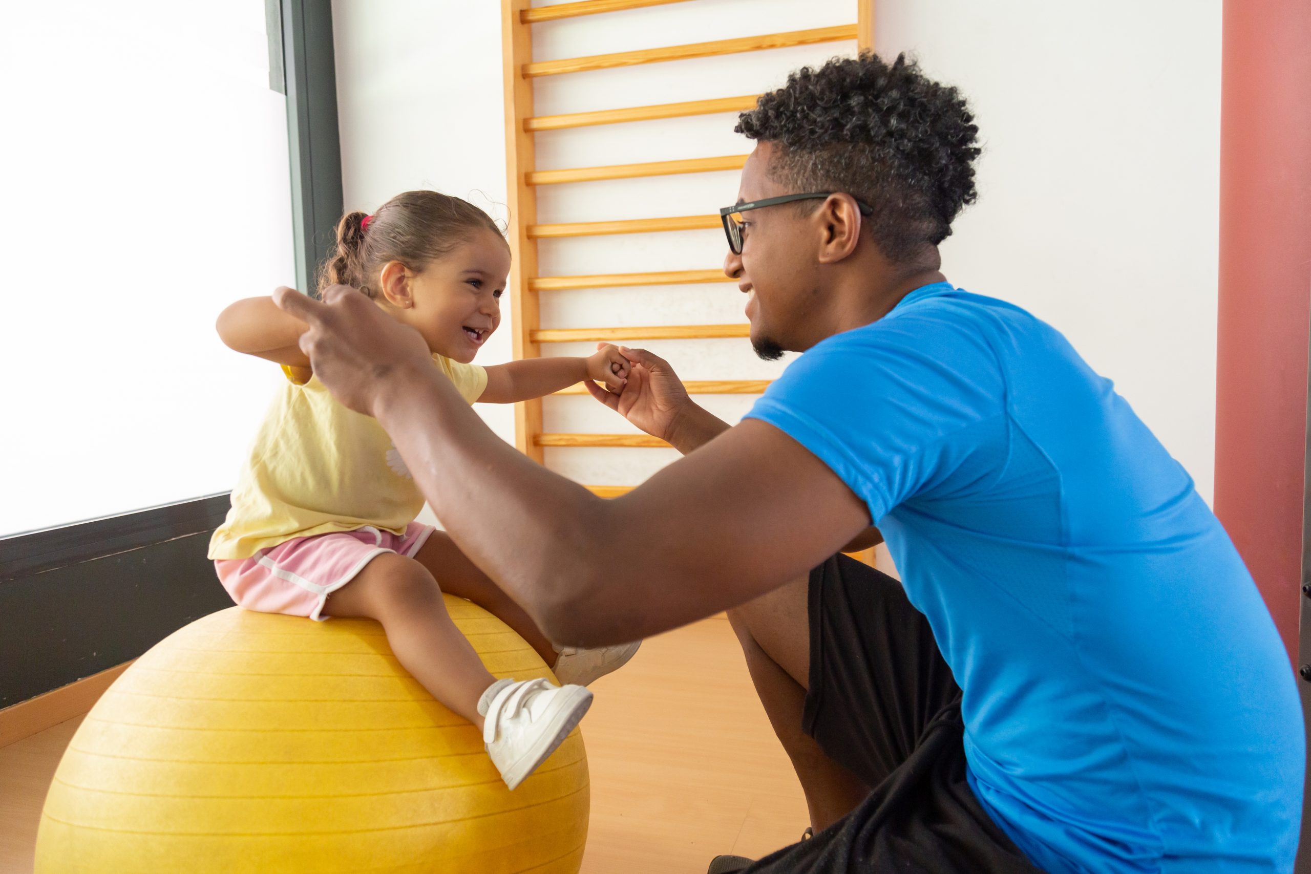Man helps a young girl balance on a yellow exercise ball in a gymnasium with a wooden climbing frame in the background.