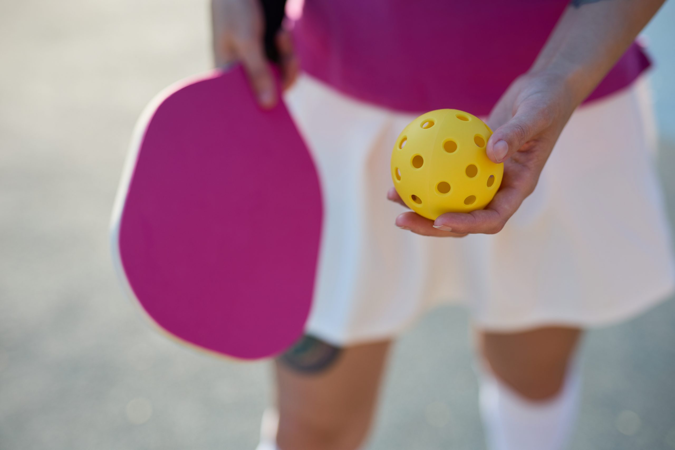 Person holding a yellow perforated pickleball and a pink paddle, dressed in a white skirt and pink top.